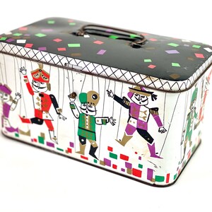 Peut inclure: Une boîte en étain vintage avec un couvercle noir et une base blanche. Le couvercle est décoré de confettis colorés. La base présente une illustration colorée de cinq marionnettes avec des cordes attachées à leurs bras et leurs jambes. Les marionnettes sont habillées de différentes couleurs et ont des expressions faciales différentes.