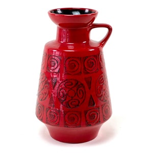 Pode incluir: Um vaso de cerâmica vermelho com uma borda preta e uma alça. O vaso tem um padrão geométrico de círculos e quadrados.