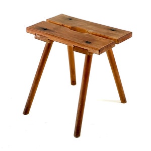 Könnte beinhalten: Ein kleiner Holzschemel mit natürlicher Oberfläche. Der Schemel hat eine rechteckige Sitzfläche aus zwei Brettern mit einem mittigen Ausschnitt. Er steht auf drei abgewinkelten, runden Beinen. Das Holz hat sichtbare Maserung und Knoten.