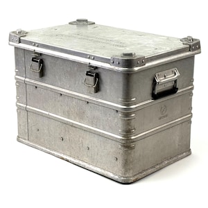 Könnte beinhalten: Eine silberne Metall-Aufbewahrungstruhe mit rechteckiger Form. Die Truhe hat einen Klappdeckel, zwei Verschlüsse und einen Griff. Die Ecken sind mit Metall verstärkt. Die Truhe besteht aus einem robusten Metallmaterial und ist für die Aufbewahrung konzipiert.