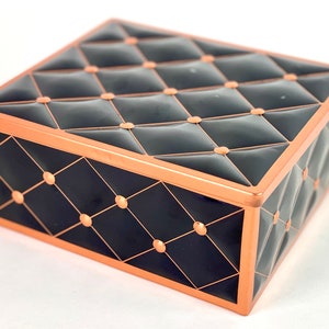 Puede incluir: Una caja rectangular negra y cobre con un diseño capitoné. La caja tiene un borde de cobre y un patrón de diamante.