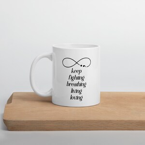 Könnte beinhalten: Weiße Keramiktasse mit einem schwarzen Unendlichkeitszeichen und dem Text "keep fighting breathing living loving".