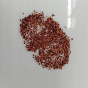 May include: A collection of small, round, reddish-brown gemstones scattered on a white surface. The gemstones are of varying sizes and appear to be faceted, reflecting light. The overall impression is of a pile of loose, raw gemstones.