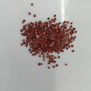 Puede incluir: Una colección de pequeñas gemas triangulares de color marrón rojizo dispersas sobre una superficie blanca. Las gemas son de forma y tamaño uniformes, con un aspecto pulido. La imagen está bien iluminada, resaltando el color y las facetas de las piedras.