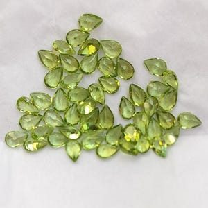 May include: A collection of pear-shaped green gemstones, also known as peridot, displayed on a white surface.