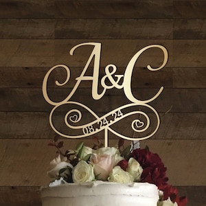 May include: A gold-coloured cake topper featuring the letters "A & C" in a script font, joined by an infinity symbol. The date "08.24.24" is inscribed within the symbol. The topper sits atop a cake adorned with white and red flowers.