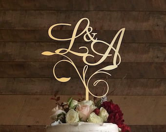 May include: A gold wedding cake topper featuring the letters L & A in a cursive script, with a decorative vine design. The topper is placed on a white cake decorated with white and pink flowers.