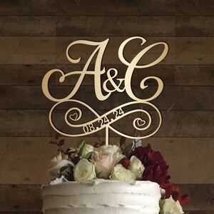 Peut inclure: Topper de gâteau de mariage en or avec les initiales A & C et la date 08.24.24. Le topper est placé sur un gâteau blanc décoré de fleurs.