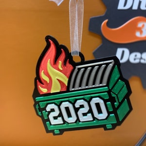 以下が含まれることがあります： 緑、黒、銀の3Dプリントされたクリスマスオーナメントで、ゴミ箱の形をしています。上部に炎が描かれています。ゴミ箱の側面には2020年が印刷されています。