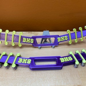 May include: Two purple and yellow face mask extenders with the letters "BHS" printed on them. The extenders have a series of yellow musical note-shaped hooks on each side. The background is a plain brown surface.