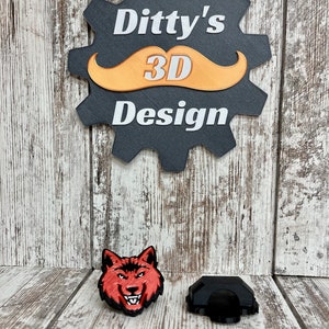 Könnte beinhalten: Ein 3D-gedruckter roter Wolfskopf und ein schwarzes 3D-gedrucktes Objekt. Der Wolfskopf hat einen grimmigen Ausdruck mit scharfen Zähnen und einer schwarzen Nase. Das schwarze Objekt ist eine geometrische Form mit abgerundeter Oberseite und flachem Boden. Die Objekte befinden sich auf einer Holzoberfläche. Das Bild zeigt auch ein Schild mit der Aufschrift "Ditty's 3D Design" mit einem stilisierten Schnurrbart.