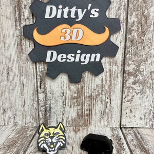 Könnte beinhalten: Ein 3D-gedruckter schwarzer und gelber Wildkatzen-Maskottchenkopf und ein schwarzes 3D-gedrucktes Objekt. Die Wildkatze hat einen weißen Mund und weiße Augen. Der Hintergrund ist eine Holzoberfläche mit einem schwarzen zahnradartigen Schild, auf dem "Ditty's 3D Design" mit einem braunen Schnurrbart steht.