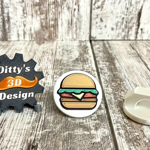 May include: An assortment of 3D-printed items. Includes a black gear-shaped logo with the text "Ditty's 3D Design" and an orange mustache, a round sticker of a burger, and a white, geometric object.