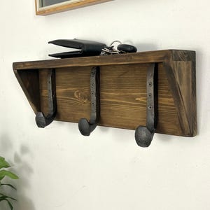 May include: A wall-mounted wooden shelf featuring a dark brown stained finish and three black metal hooks. The hooks are shaped like railroad spikes and are secured to the wood with screws. A black wallet and keys rest on the top surface.