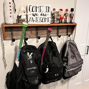 Puede incluir: Un perchero de pared de madera r&uacute;stica con ganchos de metal que sostiene tres mochilas y dos bates de b&eacute;isbol. Sobre el estante hay un letrero blanco con el texto negro COME IN we are AWESOME, flanqueado por un mu&ntilde;eco de nieve y cuatro cascanueces.
