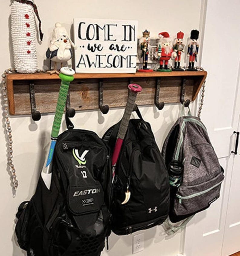 Puede incluir: Un perchero de pared de madera r&uacute;stica con ganchos de metal que sostiene tres mochilas y dos bates de b&eacute;isbol. Sobre el estante hay un letrero blanco con el texto negro COME IN we are AWESOME, flanqueado por un mu&ntilde;eco de nieve y cuatro cascanueces.