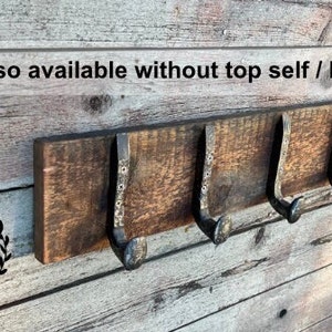 May include: A rustic wooden coat rack with three metal hooks. The rack is mounted on a wooden wall. The text "Also available without top self / borders" is visible above the rack.