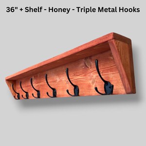 May include: A 36-inch honey-colored wooden wall shelf featuring six black metal triple hooks. The text 36" + Shelf - Honey - Triple Metal Hooks is displayed above the shelf, which has a visible wood grain and angled side supports.