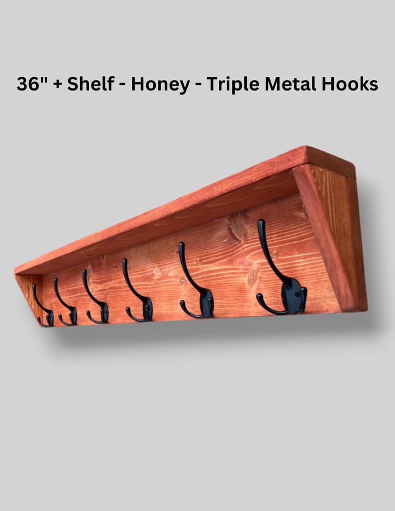 May include: A 36-inch honey-colored wooden wall shelf featuring six black metal triple hooks. The text 36" + Shelf - Honey - Triple Metal Hooks is displayed above the shelf, which has a visible wood grain and angled side supports.