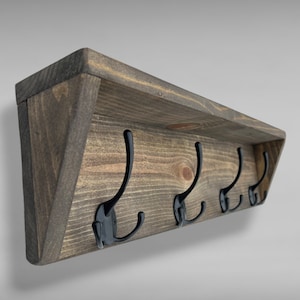 May include: A wall-mounted coat rack featuring a dark brown stained wooden shelf with a flat top and angled side supports. Four black metal double hooks are evenly spaced along the front wooden panel, each with a curved upper and lower arm.