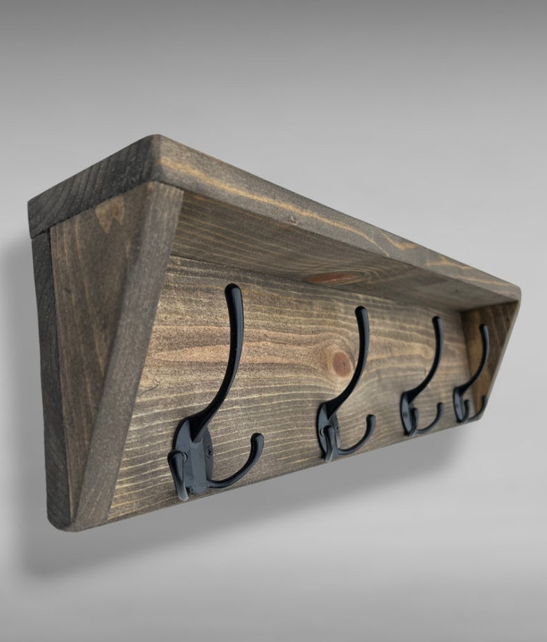 May include: A wall-mounted coat rack featuring a dark brown stained wooden shelf with a flat top and angled side supports. Four black metal double hooks are evenly spaced along the front wooden panel, each with a curved upper and lower arm.