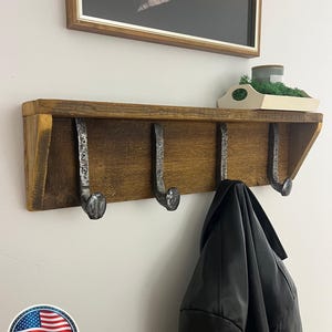Puede incluir: Un estante de madera montado en la pared con un acabado marr&oacute;n oscuro y cuatro ganchos de metal con forma de clavos de v&iacute;a f&eacute;rrea. Los ganchos est&aacute;n asegurados con tornillos visibles. Una chaqueta negra cuelga de un gancho y una bandeja.