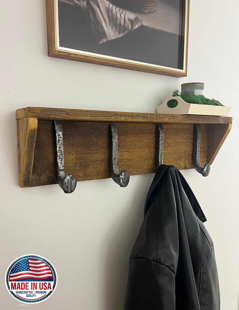 Puede incluir: Un estante de madera montado en la pared con un acabado marr&oacute;n oscuro y cuatro ganchos de metal con forma de clavos de v&iacute;a f&eacute;rrea. Los ganchos est&aacute;n asegurados con tornillos visibles. Una chaqueta negra cuelga de un gancho y una bandeja.