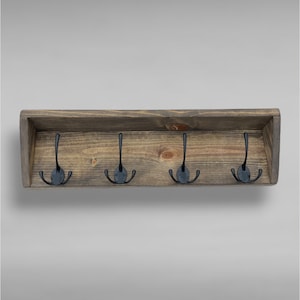 May include: A rectangular wall-mounted coat rack made of dark stained wood with a visible grain pattern. The rack features four black metal hooks, each with three prongs, securely attached to the wooden backboard within a shallow shelf frame.