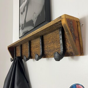 Puede incluir: Un perchero de pared construido con una pieza horizontal de madera marr&oacute;n desgastada que cuenta con cuatro ganchos de metal negro con forma de clavos de v&iacute;a f&eacute;rrea fijados con tornillos. Una chaqueta de cuero negro cuelga del gancho izquierdo.
