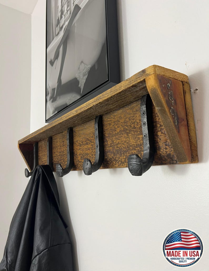 Puede incluir: Un perchero de pared construido con una pieza horizontal de madera marr&oacute;n desgastada que cuenta con cuatro ganchos de metal negro con forma de clavos de v&iacute;a f&eacute;rrea fijados con tornillos. Una chaqueta de cuero negro cuelga del gancho izquierdo.