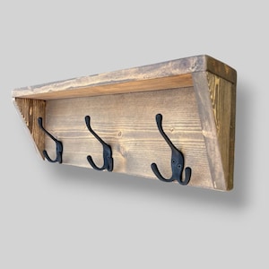 May include: A wall-mounted coat rack made of stained brown wood with a top shelf and three black metal double hooks. The wooden structure features visible grain patterns and angled side supports, providing a functional storage solution.