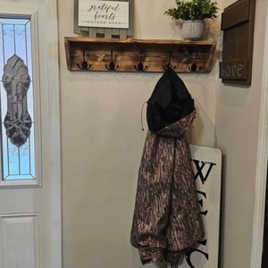 May include: A wall-mounted wooden coat rack with a dark stained finish and five black metal hooks. A camouflage-patterned jacket with a black hood hangs from one of the hooks. The rack features a flat top shelf for storage and display.