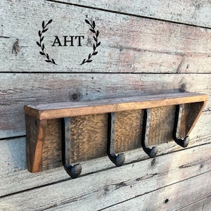 May include: A rustic wooden shelf with three metal hooks. The shelf is mounted on a wood wall. The shelf is stained a dark brown color. The hooks are black and have a hammered finish. The shelf is labeled "AHT".