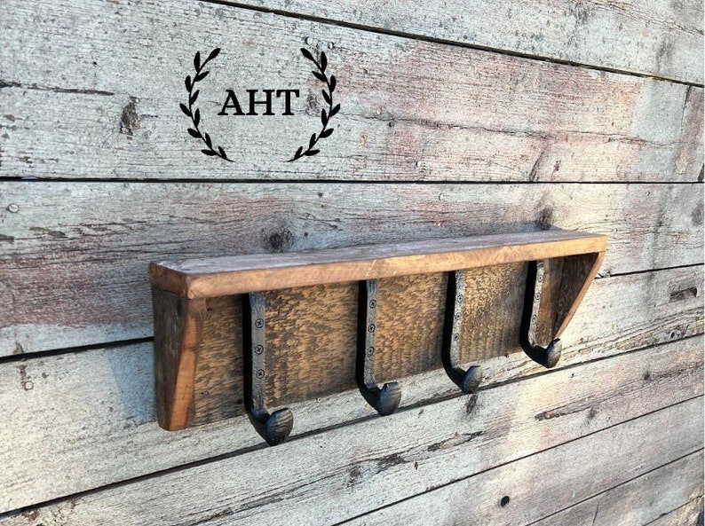 May include: A rustic wooden shelf with three metal hooks. The shelf is mounted on a wood wall. The shelf is stained a dark brown color. The hooks are black and have a hammered finish. The shelf is labeled "AHT".