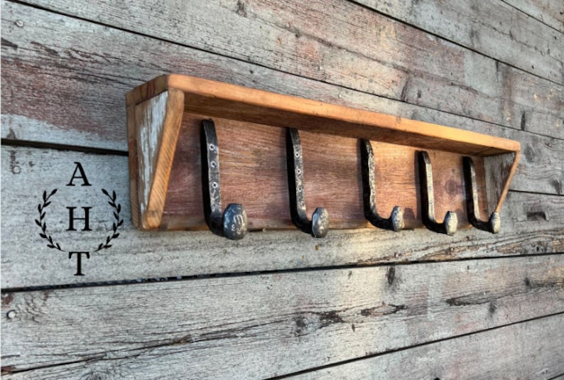 Puede incluir: Un estante de madera montado en la pared con cinco ganchos de metal oscuro en forma de clavos de v&iacute;a f&eacute;rrea. El estante tiene un acabado de veta de madera natural con tonos claros visibles. Las letras AHT est&aacute;n impresas a la izquierda en un dise&ntilde;o de laurel.