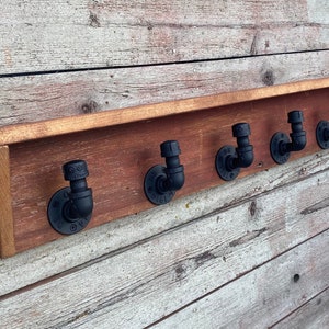 May include: A rustic wooden shelf with five black pipe hooks. The shelf is mounted on a white wood wall.