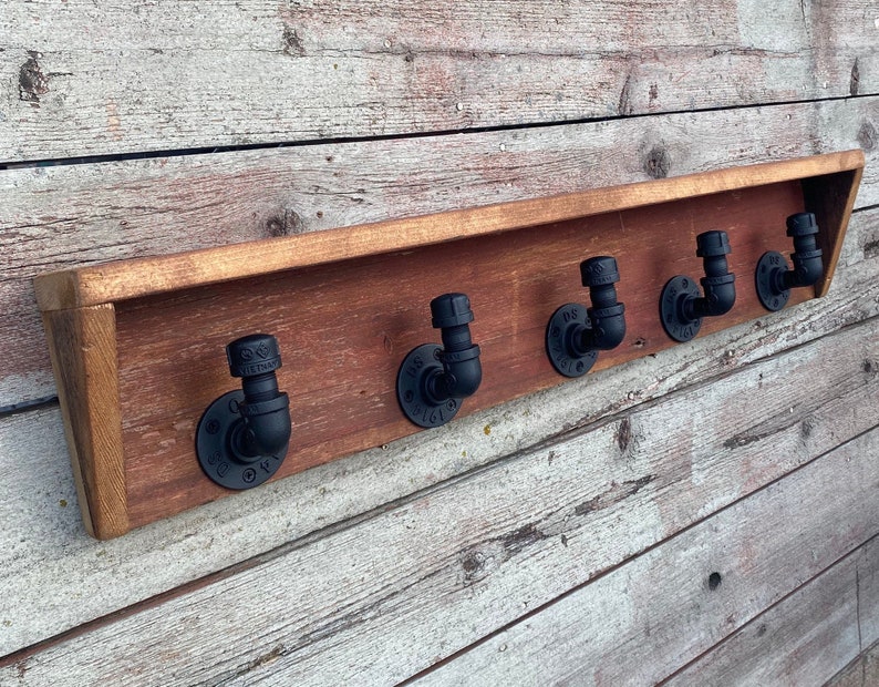 May include: A rustic wooden shelf with five black pipe hooks. The shelf is mounted on a white wood wall.