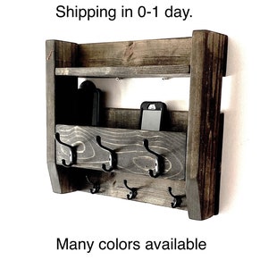 May include: A dark brown stained wooden wall organizer featuring a top shelf, a middle storage compartment, and six black metal hooks arranged in two rows. The unit has a rectangular frame with visible wood grain and vertical side supports.