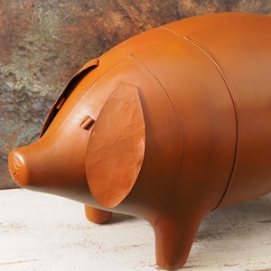 May include: A brown pig-shaped ottoman crafted from smooth material with visible stitched seams. The figure features a rounded body, four short legs, a snout, and two flat, oval-shaped ears positioned on the sides of the head.