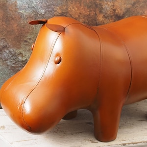 May include: A smooth, tan-colored leather ottoman shaped like a hippopotamus. The figure features stitched panel construction, small rounded ears, and circular eyes. The body is rounded with four short, sturdy legs, presenting a clean, uniform surface.