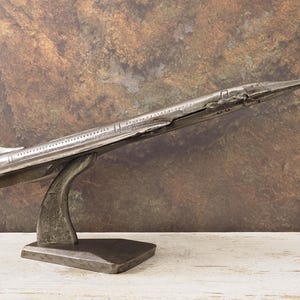 Könnte beinhalten: Ein silberfarbenes Concorde-Modellflugzeug, das auf einem geschwungenen Ständer mit rechteckiger Basis ausgestellt ist. Das Modell hat detaillierte Flügel, Heck und Rumpf und besteht aus einem metallischen Material. Der Hintergrund ist eine strukturierte, braun-beige Oberfläche.
