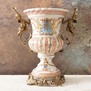 Może przedstawiać: Ozdobny, zabytkowy wazon ceramiczny o szczegółowym wzorze. Wazon ma biało-różowy korpus ze złotymi akcentami, w tym figurami cherubinów trzymających trąbki. Podstawa i uchwyty są również złote, co dodaje mu antycznego wyglądu.