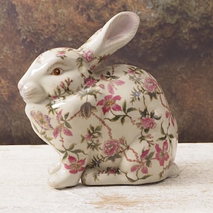 Op de afbeelding: Een keramieken konijnfiguur met een bloemmotief in roze, groen en paars op een crèmekleurige achtergrond. Het konijn heeft bruine ogen en een roze binnenkant van de oren. De figuur staat op een wit oppervlak.