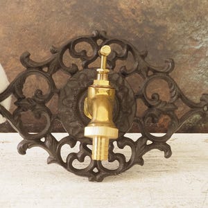 Może przedstawiać: Ozdobny, ciemnobrązowy kran ścienny z żeliwa z mosiężnym kranem. Kran jest wyśrodkowany na dekoracyjnej, ażurowej płycie tylnej. Tło to rozmyta mieszanka brązu i zieleni.