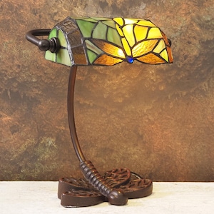 May include: A stained glass desk lamp with a dragonfly design. The lamp has a bronze-coloured metal base and a curved stem. The lampshade is made of yellow, green, and orange glass pieces, with a blue accent. The lamp is designed to sit on a desk or table.