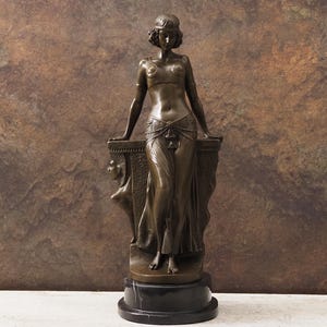 May include: Bronze statue of a woman in a flowing gown, leaning on a decorative pedestal. The statue has a dark brown patina and stands on a black marble base. The woman's arms are resting on the pedestal, and a carved figure is visible on the side.
