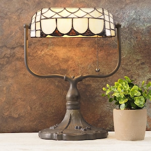 May include: A vintage-style desk lamp with a stained glass shade. The shade features a cream and brown pattern, and the lamp has a bronze-colored metal base. A small potted plant sits to the right of the lamp.