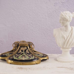 Könnte beinhalten: Ein gold- und schwarzes Tintenfass mit Art-Nouveau-Design. Das Tintenfass hat einen Deckel und ist mit grünen und goldenen floralen Motiven verziert. Das Tintenfass steht auf einer weißen Oberfläche neben einer weißen Gipsbüste einer Frau.