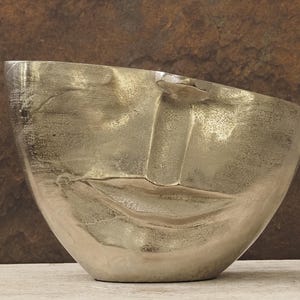 May include: A silver-toned bowl with a face design. The bowl has a smooth, reflective surface with a sculpted face, including lips and a nose. The bowl is set against a brown and beige background.