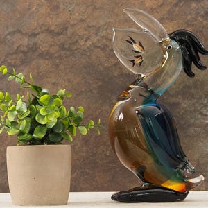 Könnte beinhalten: Eine dekorative Glaspelikan-Skulptur in Braun, Blau und Schwarz. Der Schnabel des Pelikans ist geöffnet und zeigt zwei kleine Fische. Links von der Skulptur befindet sich eine kleine Topfpflanze mit grünen Blättern.